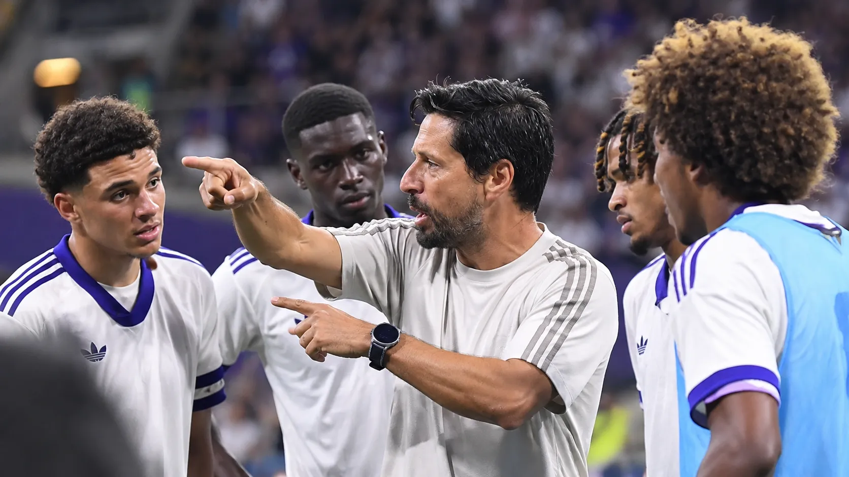 Een coach geeft aanwijzingen aan spelers langs de zijlijn van een voetbalstadion.