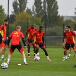 Een groep spelers en stafleden in rode trainingskleding tijdens een oefensessie op een trainingsveld.