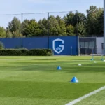 Leeg trainingsveld van KRC Genk met loopoefeningen uitgezet met kegels en een clubbord langs de zijlijn.