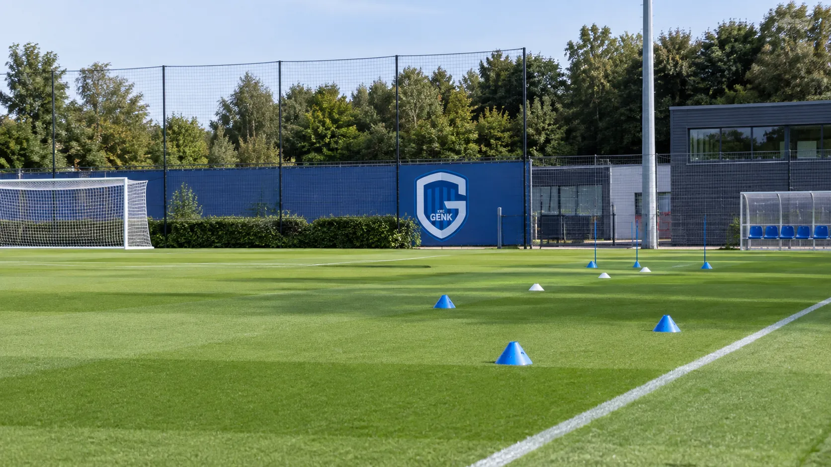 Leeg trainingsveld van KRC Genk met loopoefeningen uitgezet met kegels en een clubbord langs de zijlijn.