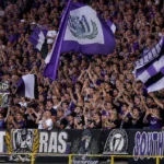 Een volle groep RSC Anderlecht-supporters applaudisseert in een paars-witte thuistribune.