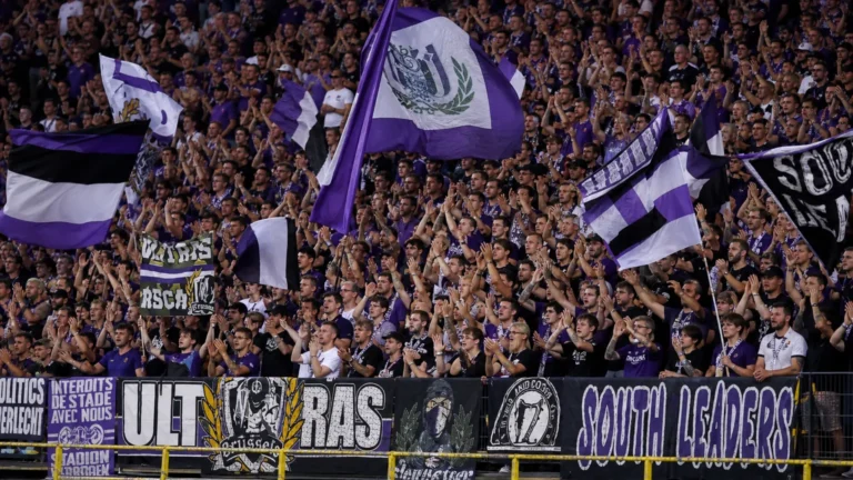Een volle groep RSC Anderlecht-supporters applaudisseert in een paars-witte thuistribune.