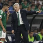 Een coach in donker pak geeft langs de zijlijn aanwijzingen aan een speler in groen tenue.