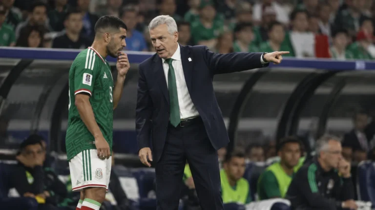 Een coach in donker pak geeft langs de zijlijn aanwijzingen aan een speler in groen tenue.