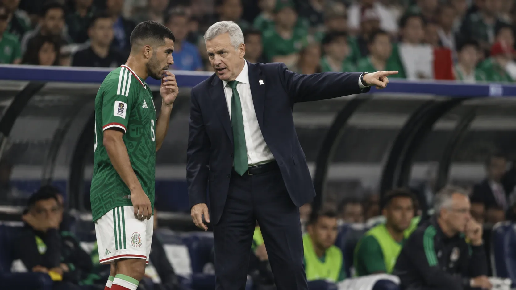 Een coach in donker pak geeft langs de zijlijn aanwijzingen aan een speler in groen tenue.