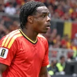 Een geconcentreerde speler in een rood Belgisch nationaal tenue op een voetbalveld.