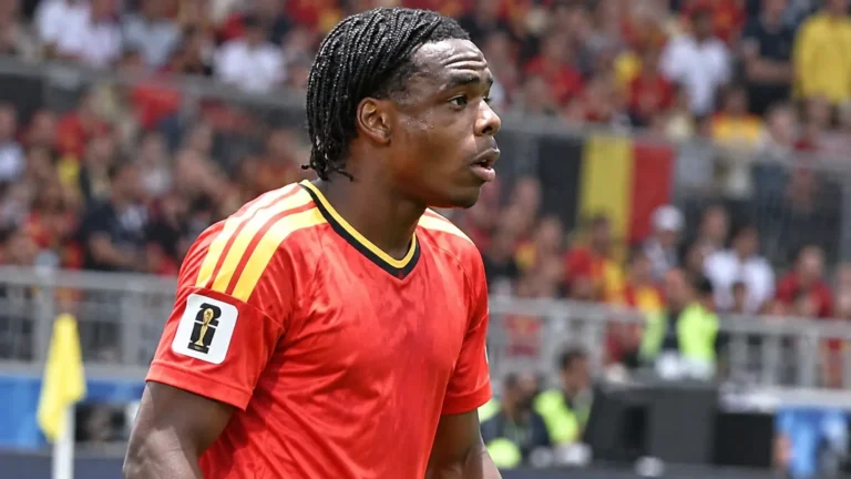 Een geconcentreerde speler in een rood Belgisch nationaal tenue op een voetbalveld.