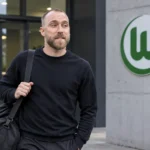 Christian Eriksen in neutrale reiskleding bij een Wolfsburg-clubgebouw.