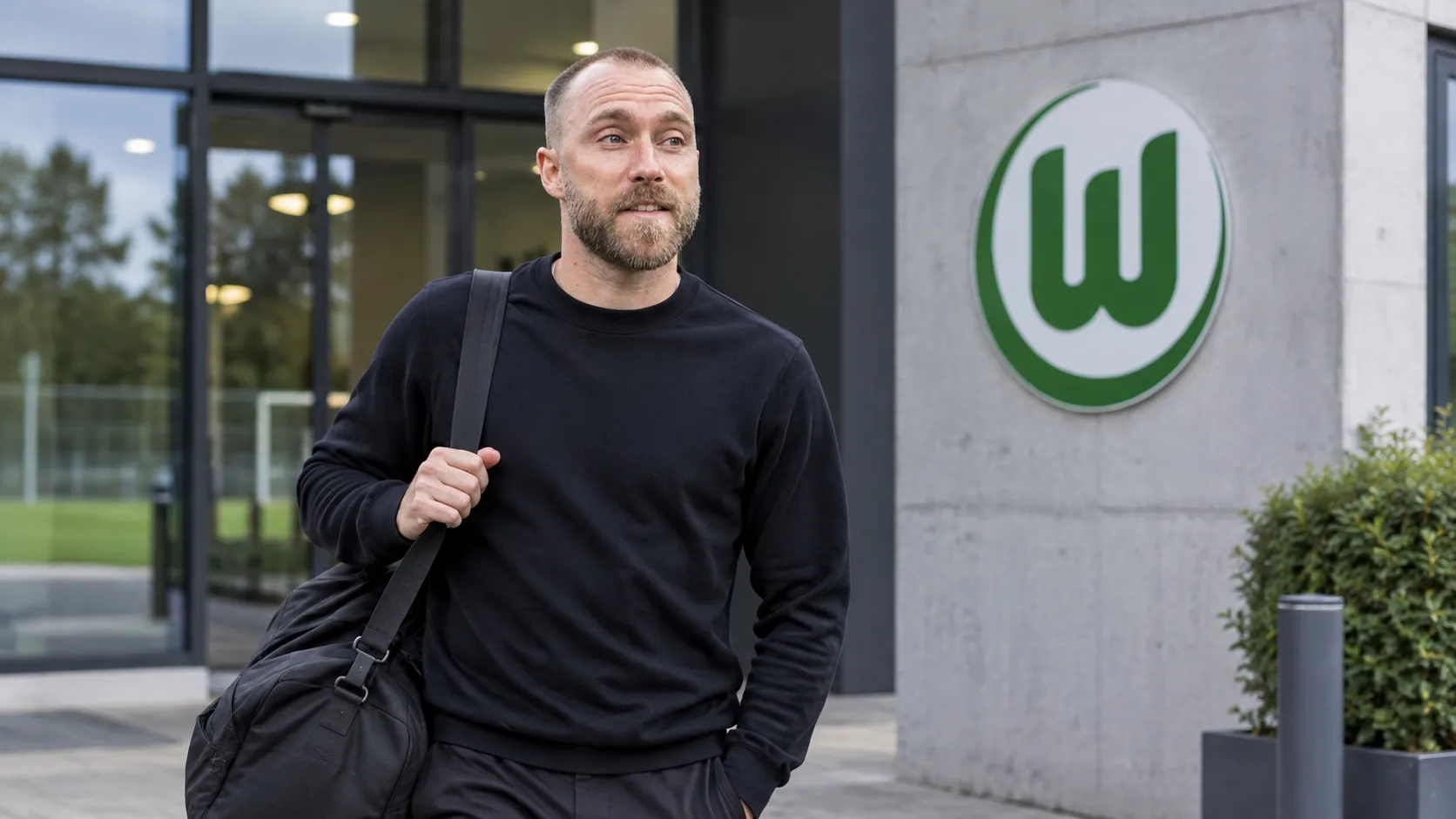 Christian Eriksen in neutrale reiskleding bij een Wolfsburg-clubgebouw.