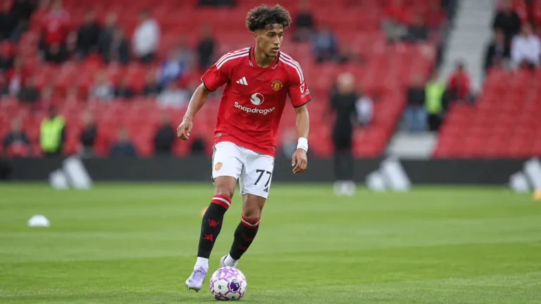 Een jonge voetballer in een rood shirt en witte broek op het veld in een stadion.