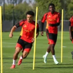 Een groep voetballers in rode trainingskleding werkt een oefening met palen af op een grasveld.