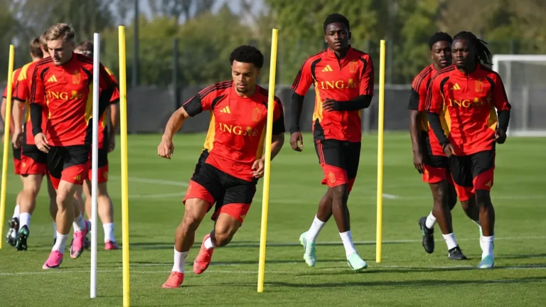 Een groep voetballers in rode trainingskleding werkt een oefening met palen af op een grasveld.