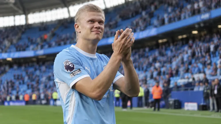 Een voetballer in een lichtblauw wedstrijdshirt applaudisseert aan de rand van het veld.