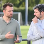 Twee niet-spelende voetbalmedewerkers in gesprek naast een trainingsveld.