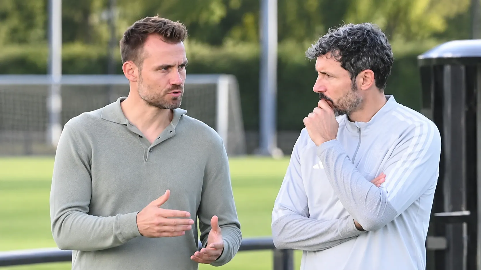 Twee niet-spelende voetbalmedewerkers in gesprek naast een trainingsveld.