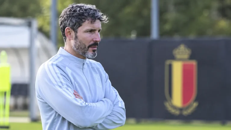 Mark van Bommel kijkt toe op het trainingsveld in Tubeke, met een vast Rode Duivels-bord op de achtergrond.