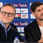Twee clubvertegenwoordigers van KRC Genk voor een blauw sponsorbord.