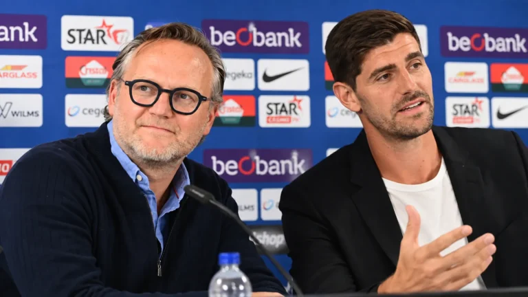 Twee clubvertegenwoordigers van KRC Genk voor een blauw sponsorbord.