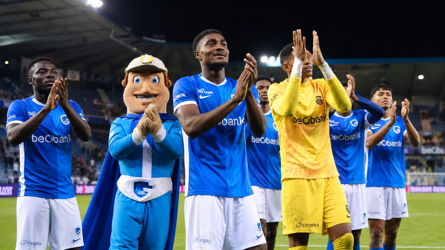 Spelers van KRC Genk en de clubmascotte applaudisseren langs het veld voor de supporters.