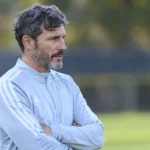 Een Belgische stafmedewerker in een lichte trainingsjas langs een trainingsveld.