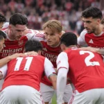Een groep spelers in rood-witte tenues vormt een huddle op het veld.