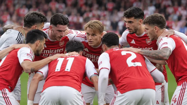 Een groep spelers in rood-witte tenues vormt een huddle op het veld.