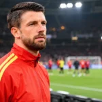 Brandon Mechele in een rode trainingsjas aan de rand van het veld in een stadion.