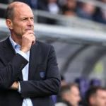 Een coach in donker pak kijkt geconcentreerd vanuit de technische zone naar het veld.