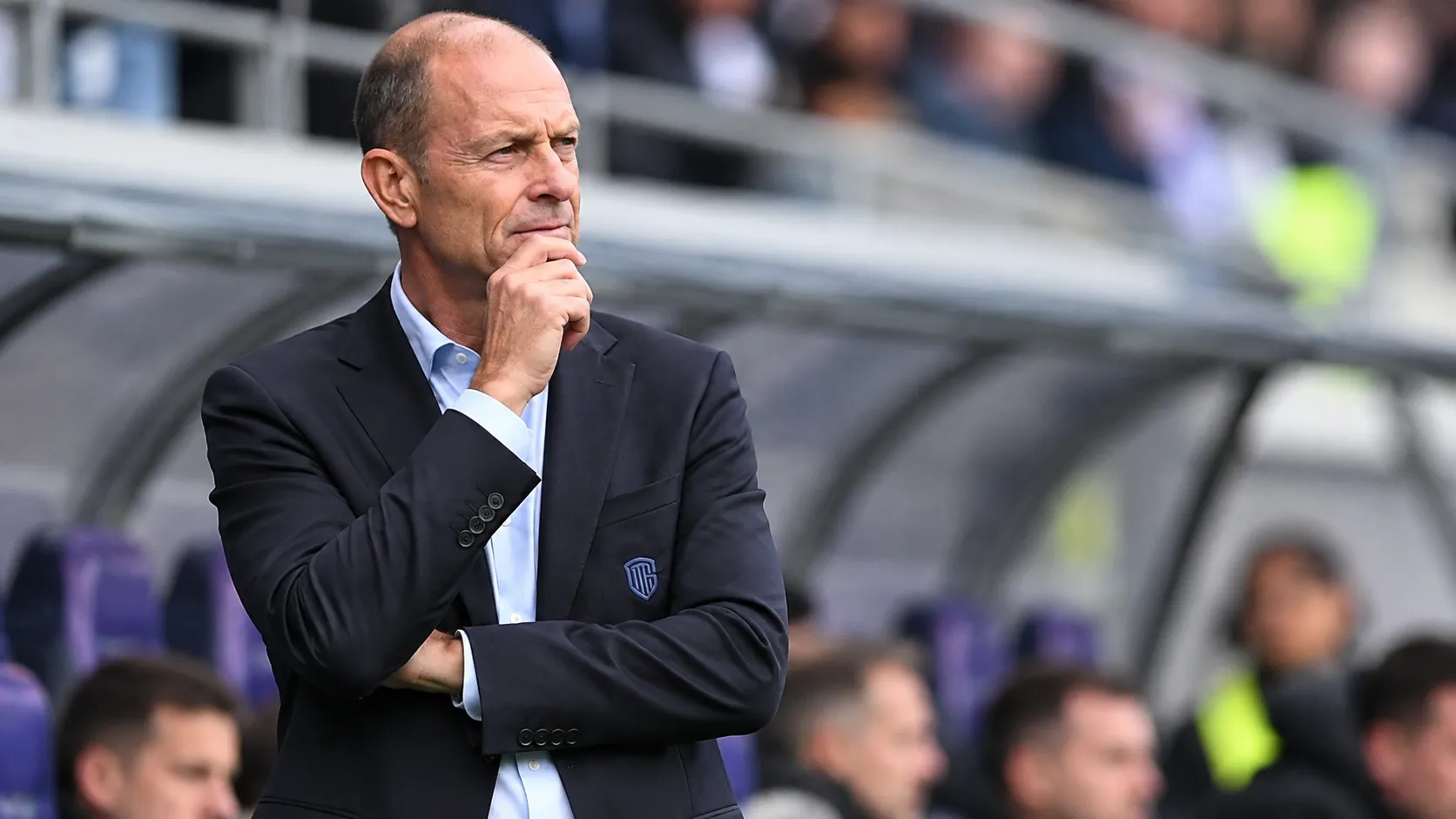 Een coach in donker pak kijkt geconcentreerd vanuit de technische zone naar het veld.