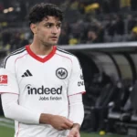 Mahmoud Dahoud in een wit wedstrijdshirt aan de zijlijn van een voetbalstadion.