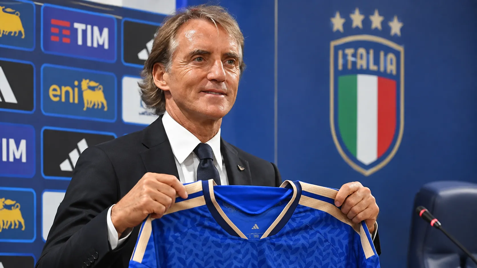 Roberto Mancini houdt een blauw Italië-shirt vast tijdens een persmoment.