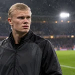 Erling Haaland in een zwarte trainingsjas aan de rand van een verlicht voetbalveld.