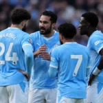 Een groep Manchester City-spelers overlegt op het veld in lichtblauwe wedstrijdtenues.