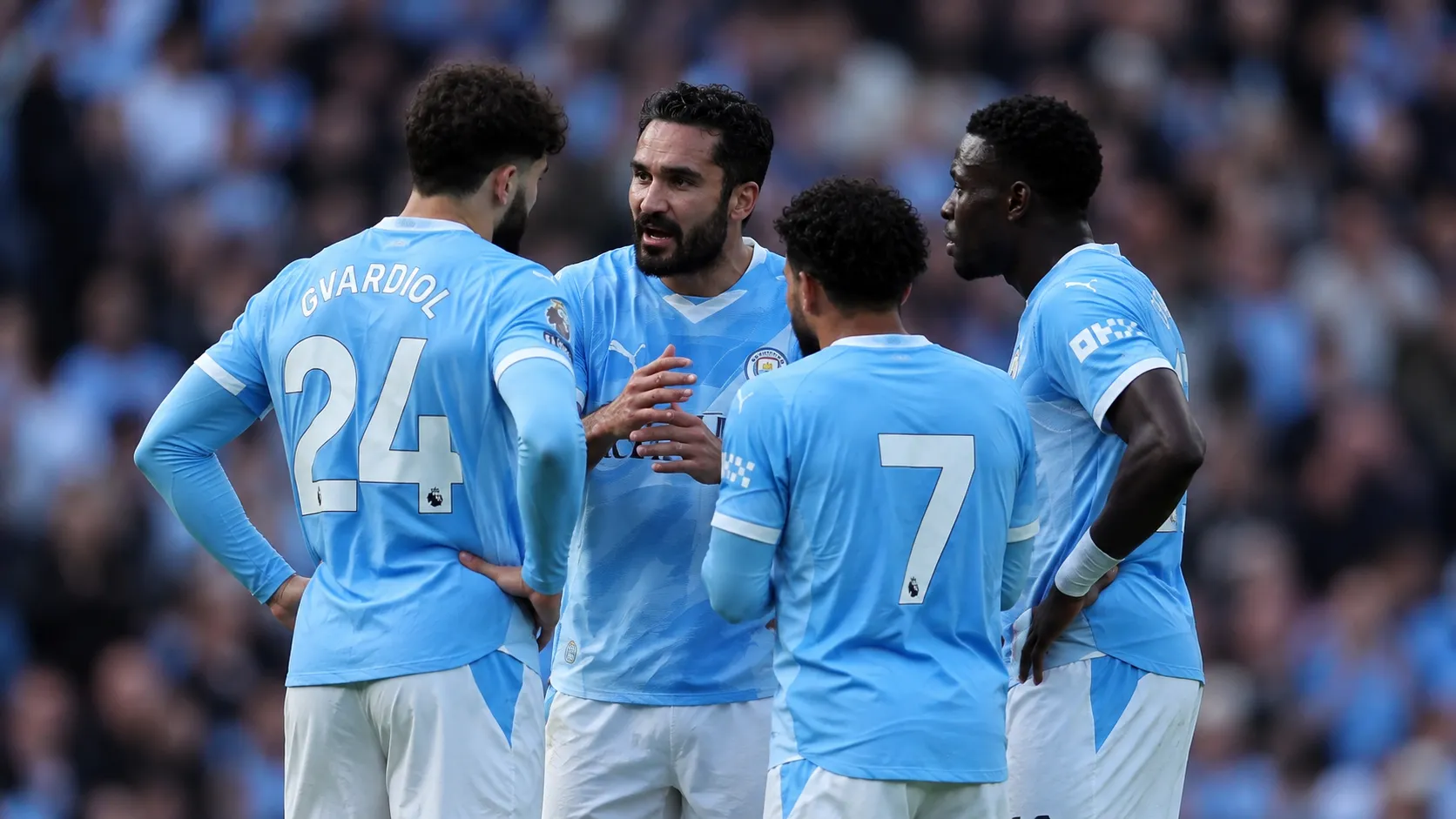 Een groep Manchester City-spelers overlegt op het veld in lichtblauwe wedstrijdtenues.