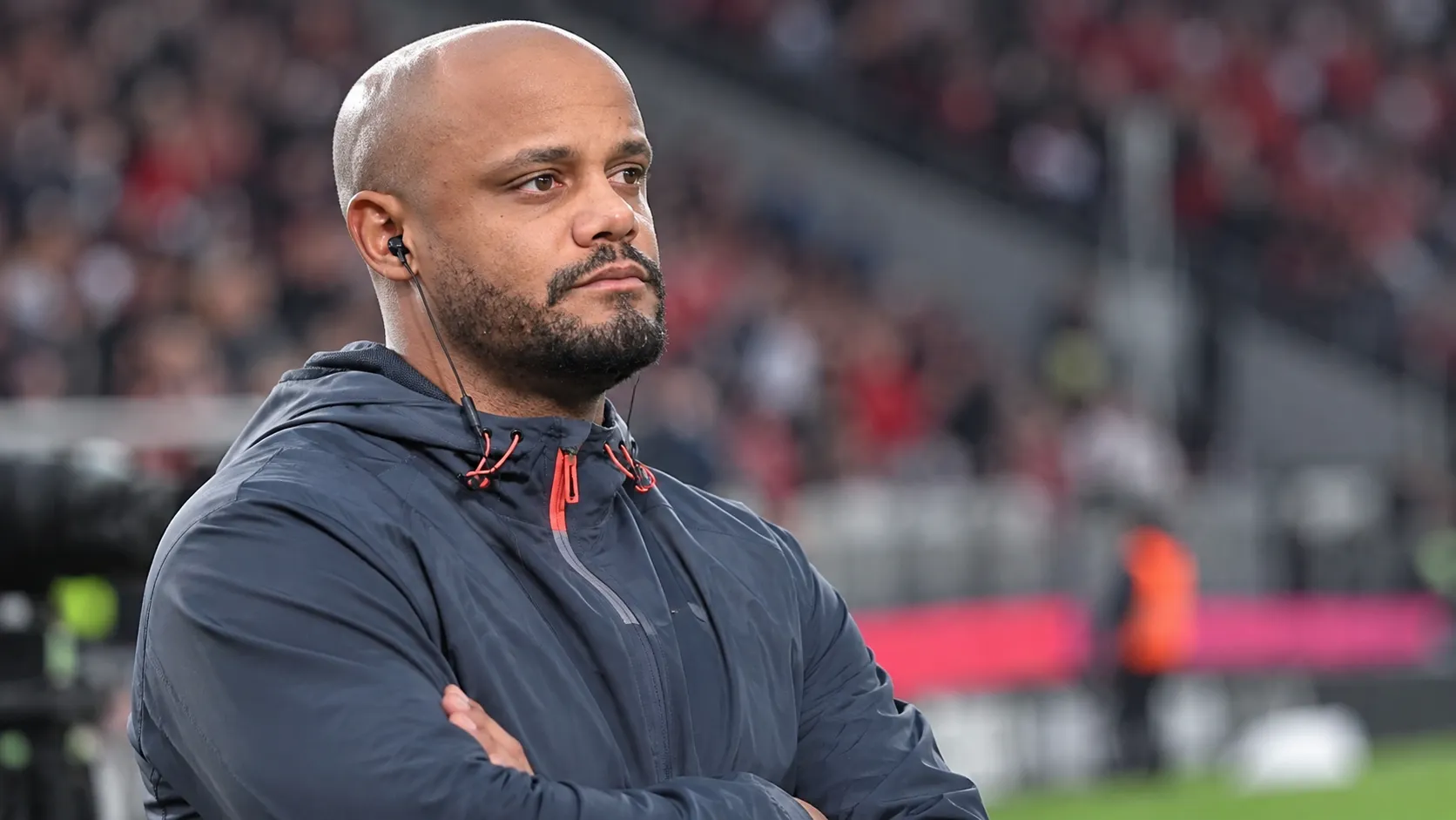 Een Bayern-medewerker in donkere sportkleding met oortje in een stadionomgeving.