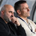 Twee niet-spelende voetbalvertegenwoordigers bekijken aandachtig het veld vanuit een overdekte dug-out.