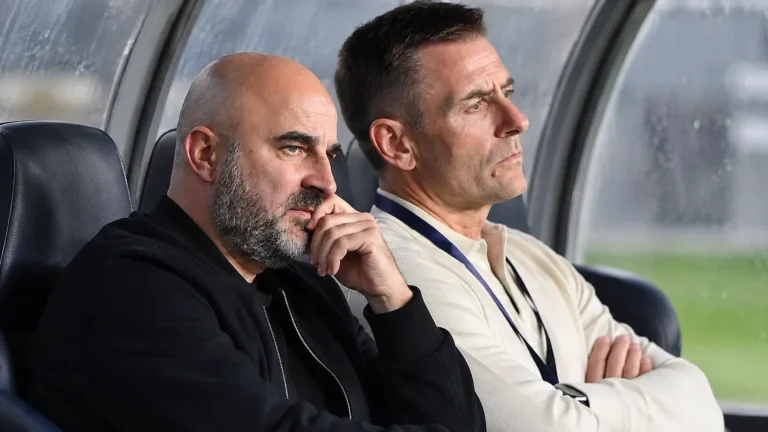 Twee niet-spelende voetbalvertegenwoordigers bekijken aandachtig het veld vanuit een overdekte dug-out.