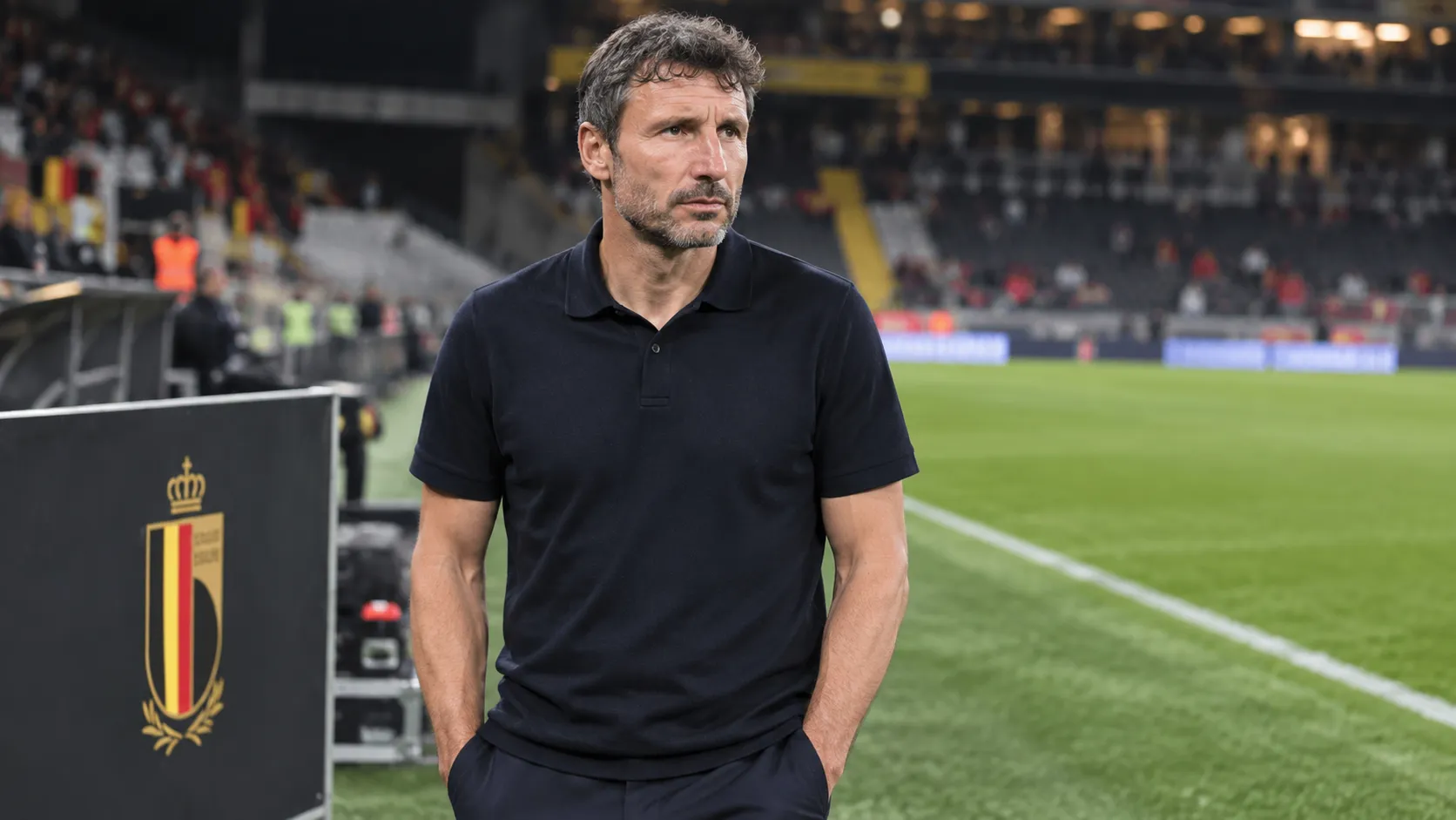 Mark van Bommel staat rustig op de zijlijn van een stadion naast een Belgisch bondslogo.
