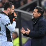 Een trainer praat langs het veld met een doelman in een lichtblauw tenue.