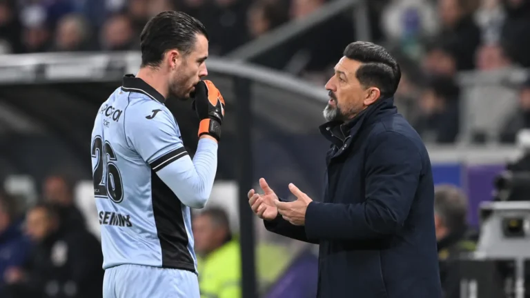 Een trainer praat langs het veld met een doelman in een lichtblauw tenue.