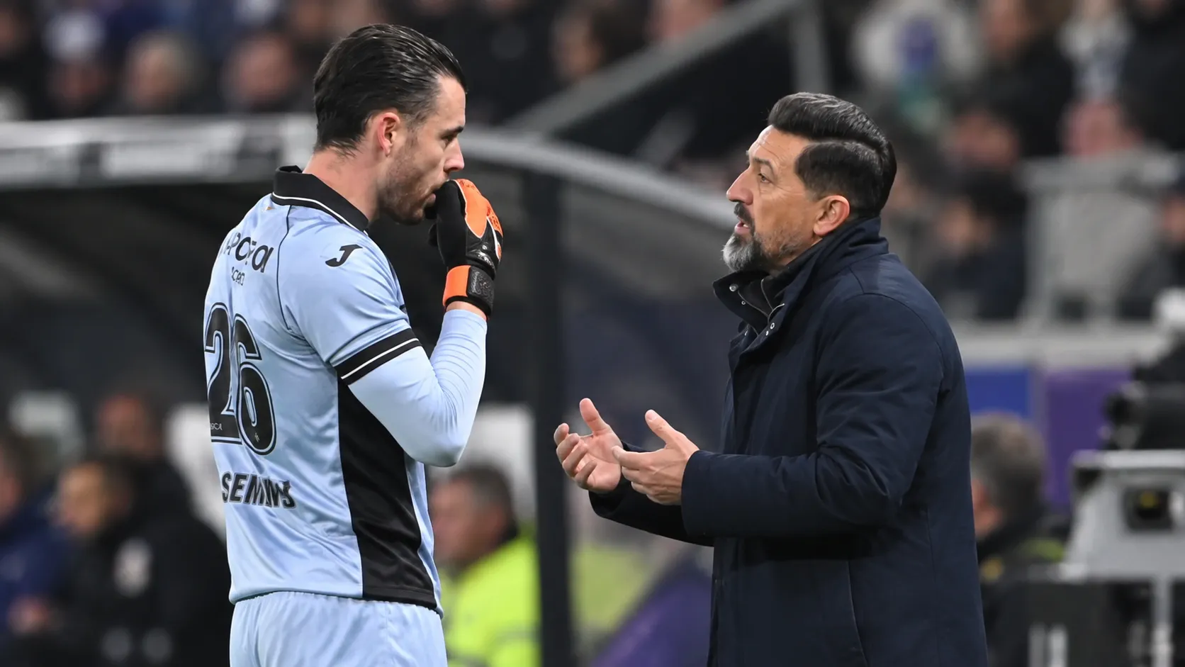 Een trainer praat langs het veld met een doelman in een lichtblauw tenue.