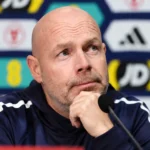 Brian Riemer spreekt op een persconferentie van Denemarken in een trainingsjas.