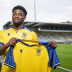 Nathanaël Mbuku poseert met een STVV-shirt tijdens een clubmediapresentatie aan het veld.