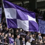 Een groep supporters met paarse en witte vlaggen op de tribune.