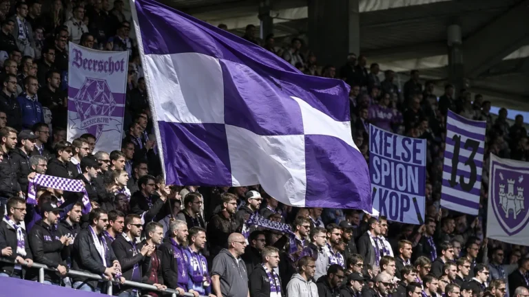 Een groep supporters met paarse en witte vlaggen op de tribune.
