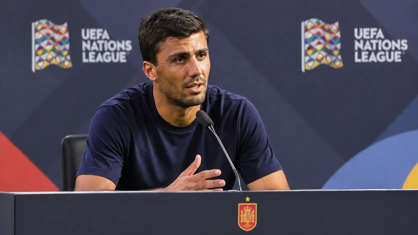 Rodri spreekt op een persconferentie achter een spreekgestoelte met een klein embleem van Spanje.