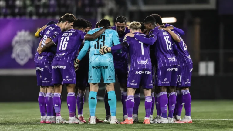 Een groep voetballers in paarse wedstrijdkleding vormt een hechte huddle op kunstgras.