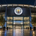 De ingang van een stadiongebouw van Manchester City met het clubembleem aan de gevel.