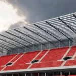 Lege tribune met rode zitjes onder de stalen dakconstructie van een stadion.