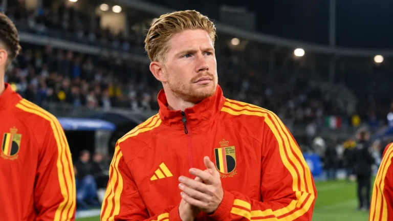 Kevin De Bruyne in een rood trainingsjack van België aan de zijlijn van een stadion.